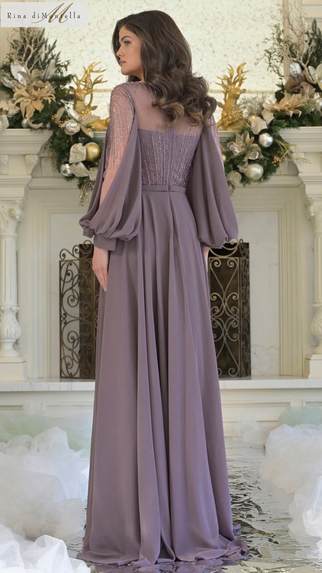 A woman stands indoors facing away in front of a white fireplace adorned with gold and white ornaments, wearing the Rina Di Montella RD3374 long formal dress in mauve chiffon with flowing sleeves, lace accents, and an elegant a-line skirt.